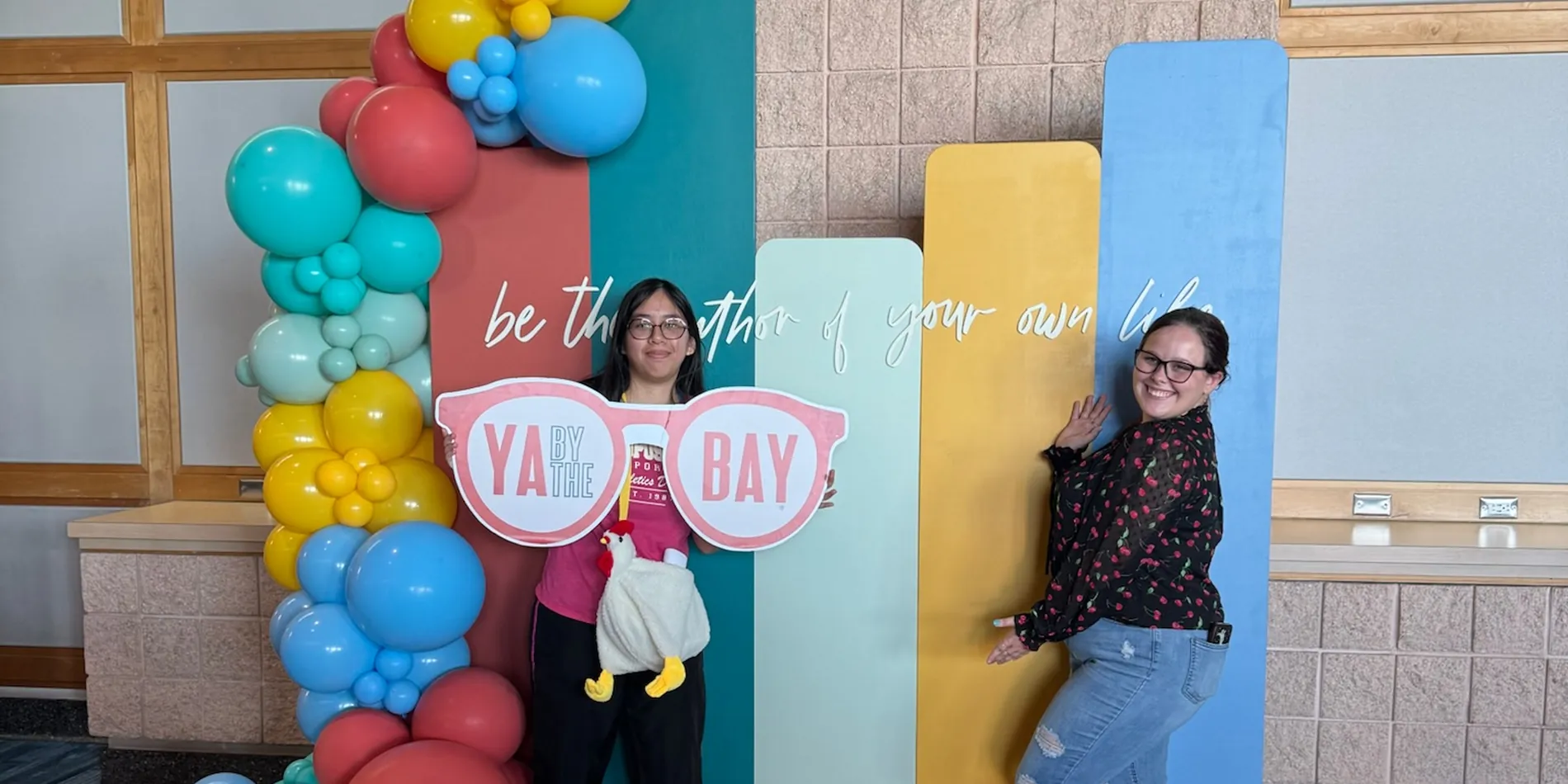 Two students holding the YA By The Bay glasses sign at the balloon backdrop